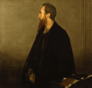 Charles De Sousy Ricketts is wearing a black cloak, only being able to see half his face and red beard that comes down to his collar bones. He is looking to his right, with hands holding one another under his cloak, beside a table.