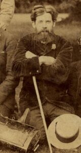 Captured image of middle-aged man (Geddes) sitting outdoors with arms crossed. There is a white hat placed at the bottom right of the image, below Patrick Geddes. Image is in sepia-tinted, light brown colour.