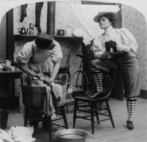 black and white photo of a woman standing with leg up on chair as a man does laundry next to her