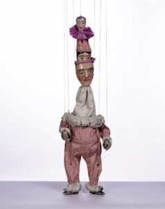 A marionette with red and white striped clothing and a long neck with three faces on top of each other.